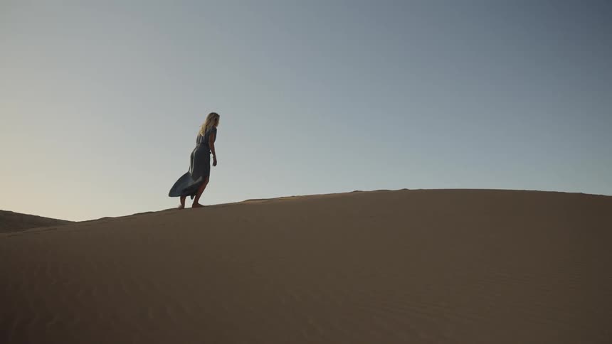 A woman walks alone up a sand dune in the desert during dawn or dusk under a clear sky