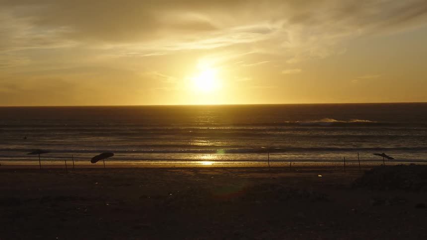 The sun sets over a calm ocean as waves gently roll toward a quiet silhouetted beach