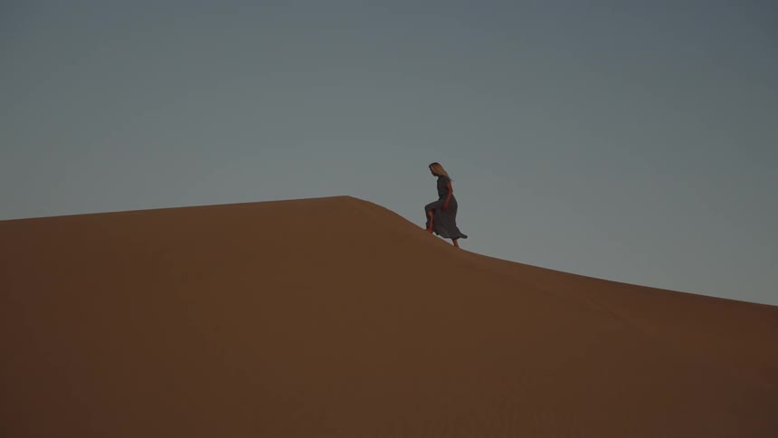 Woman wearing a long dress walks up a sand dune hill