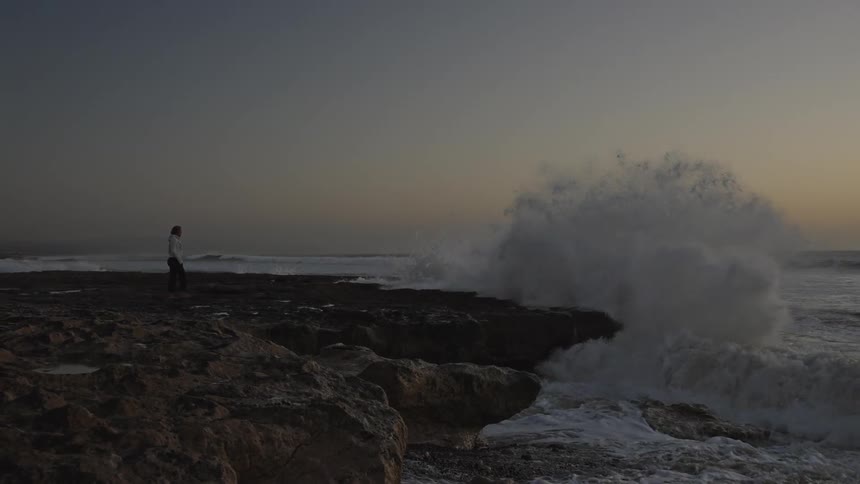 Woman walking along the coast as waves crash against rocks and splash high into the air