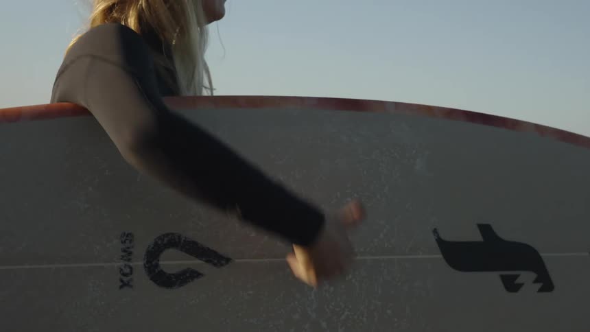 A young woman picks up her surfboard on a wet sandy beach and carries it down the shore