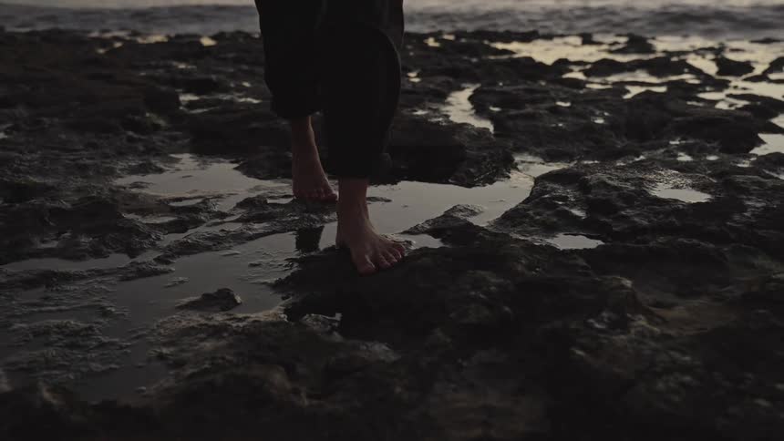 A person walks barefoot on wet jagged coastal rocks at dusk
