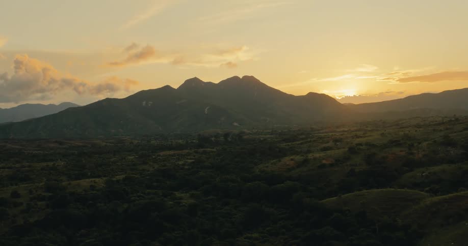 Trees and vegetation across the rural land as the sun shines through the clouds behind a mountain range