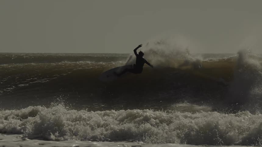 A silhouetted surfer rides a wave carving across its face as spray erupts behind them under an overcast sky