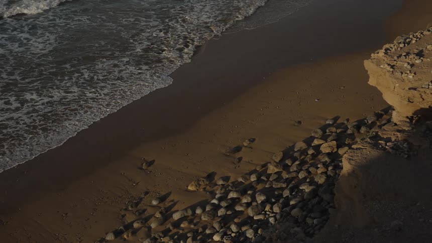 Aerial shot of waves softly washing onto a rocky sandy shoreline highlighting the peaceful contrast between land and sea