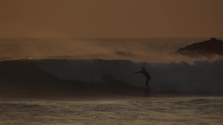 Silhouetted surfers ride waves as the sun sets casting a warm orange glow over the ocean
