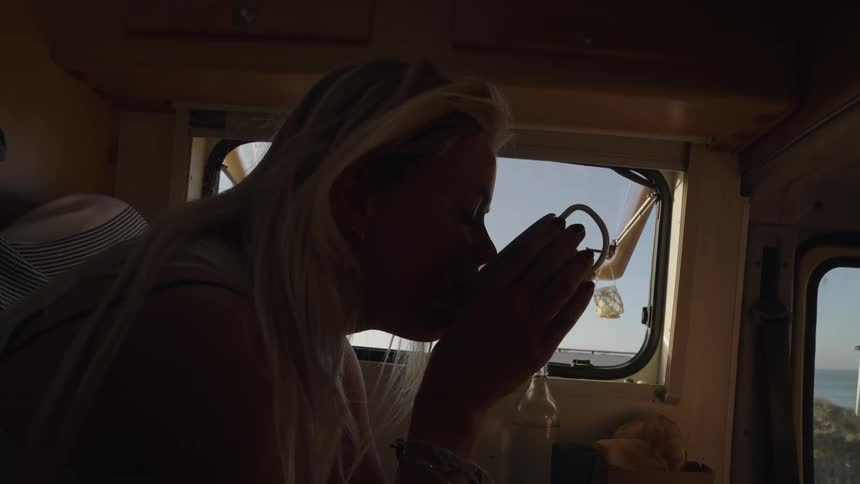 A woman drinks from a mug inside a van with sunlight streaming through the open window