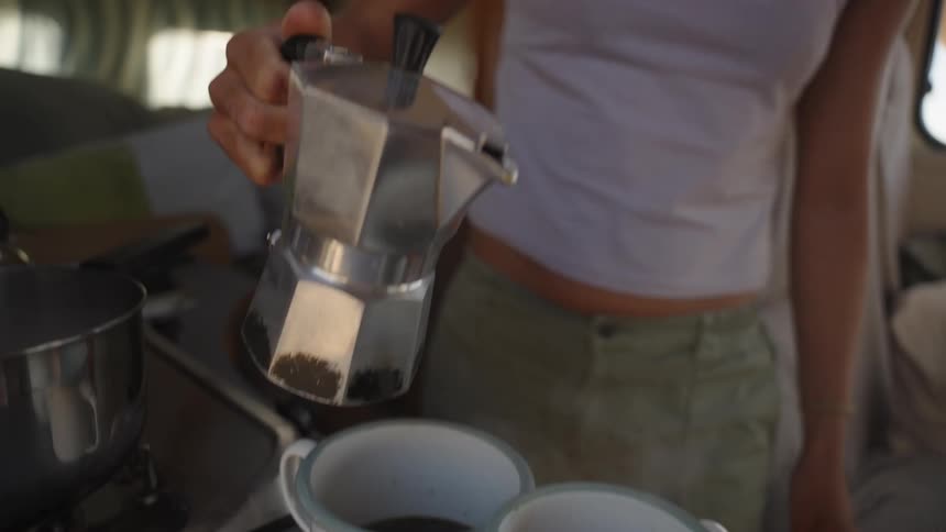 A person is pouring freshly brewed coffee from a moka pot into mugs inside a cozy sunlit camper or van