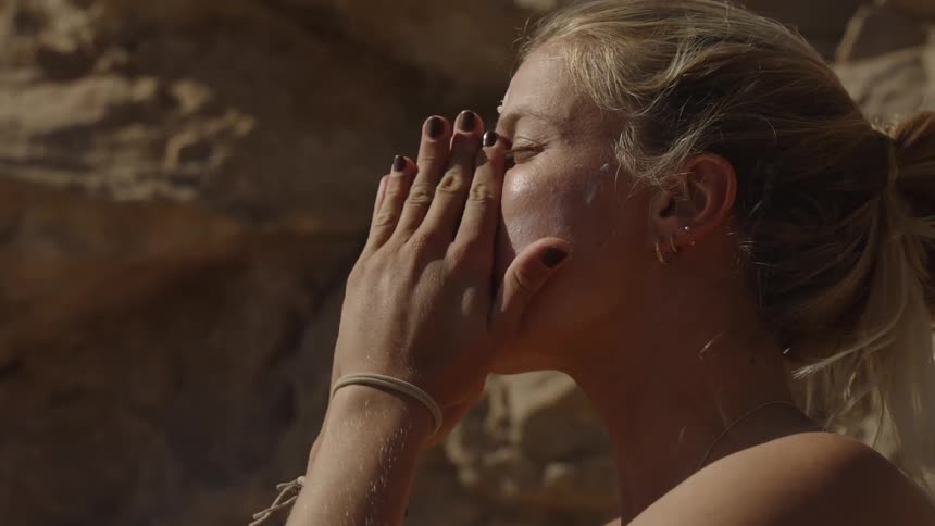A woman is seen applying sunscreen to her face under bright sunlight in a dry rocky desert environment