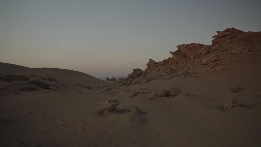 A barren desert landscape under twilight with sand dunes and rocky outcrops in view