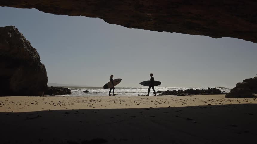 View from a cave of two people carrying surfboards on the beach as waves roll in to shore