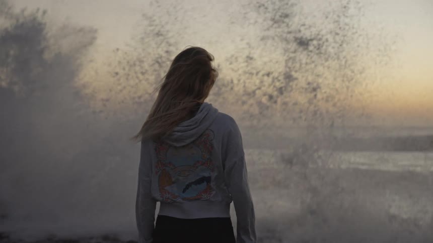 A woman faces crashing ocean waves at sunset her hair blowing in the wind