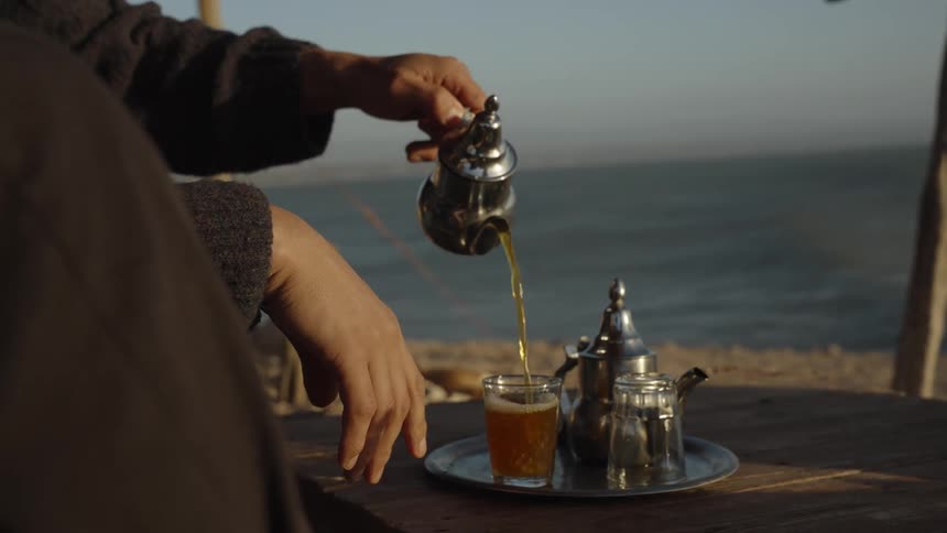 A man pouring tea from a kettle into a glass while seating on the beach