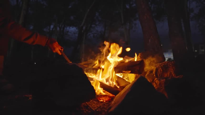 A person tends a crackling campfire at night in a wooded area