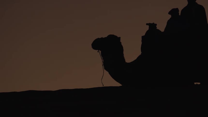 Silhouetted figures ride atop a camel resting in the desert against a dim featureless sky at either dusk or dawn