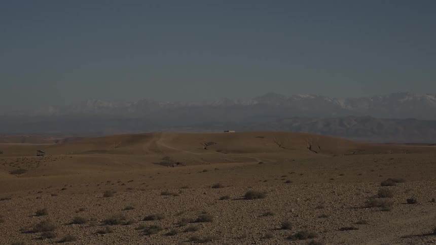 A wide landscape shot of an arid desert with scattered brush distant hills and a snow capped mountain range in the background under a clear sky