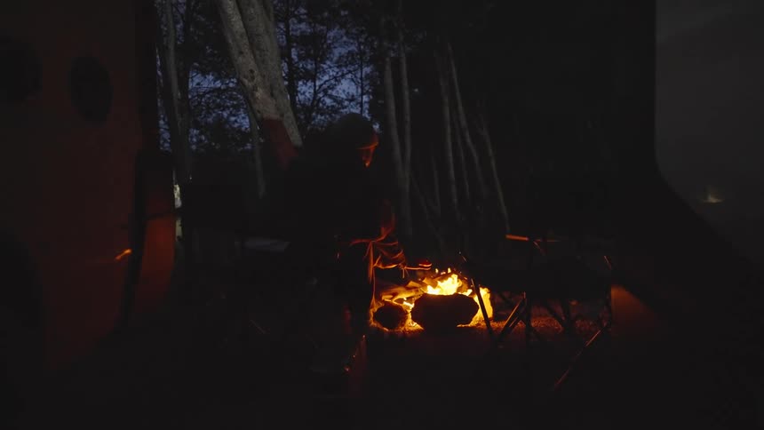 A woman takes a sit in a camping chair next to a campfire and warms up her hands