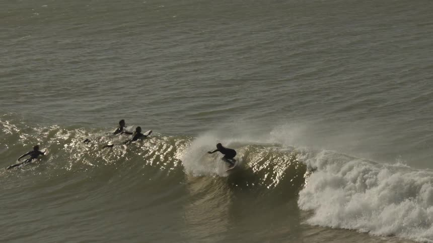 A surfer rides a powerful wave near the shore while several others wait in the water under a cloudy sky