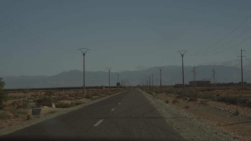 View through a dashboard of a vehicle moving along a paved road surrounded by desert vegetation and mountains in the background