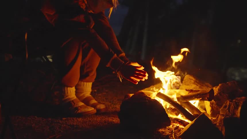 A woman in warm clothes sits by a campfire at night warming her hands as the fire glows in the forest