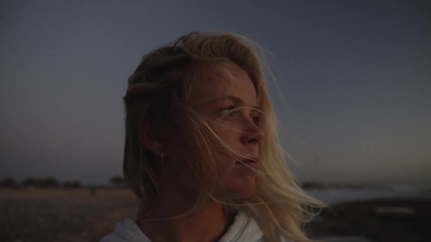 A woman with long blonde hair stands near the coastline at dusk her hair flowing in the wind as she gazes toward the horizon