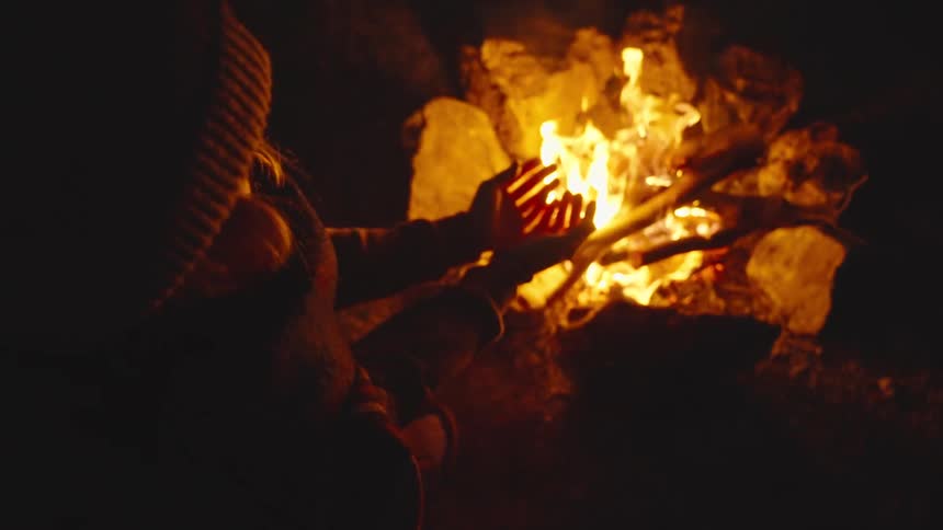 A person warms their hands by a campfire at night creating a cozy intimate outdoor moment