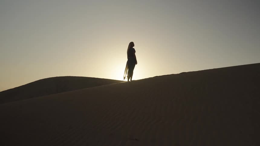 A silhouetted woman stands on a sand dune at sunset evoking a calm and contemplative mood