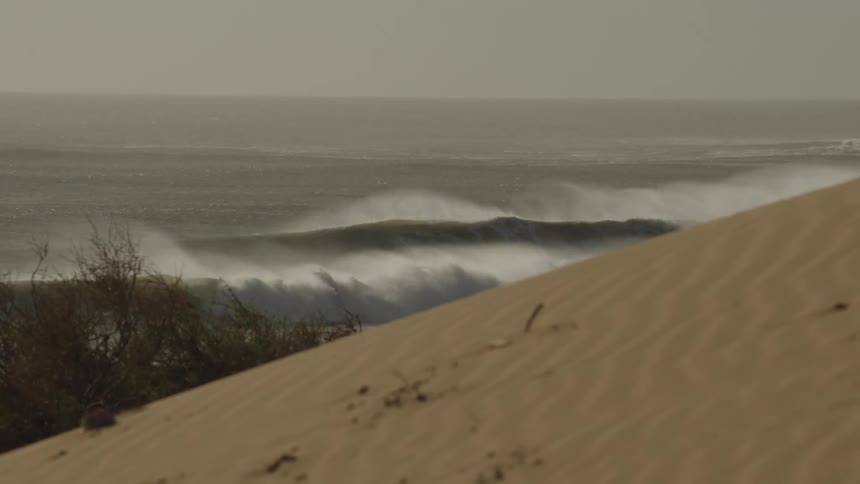 Waves crash along a coastline as seen from behind a sandy dune with sparse vegetation under a hazy sky