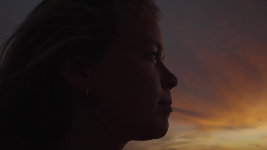 The face of a young woman her hair is moving in the wind and she is looking at the horizon where the sky is orange in the morning