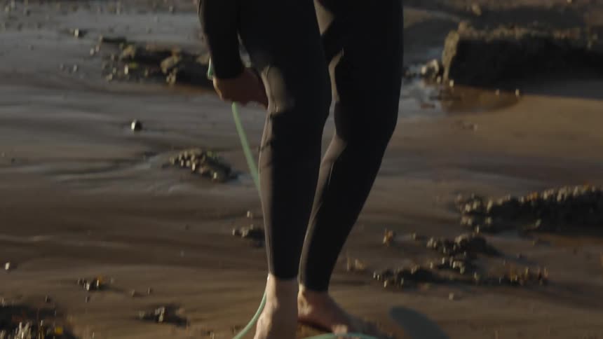 Female surfer wearing a wet suit while standing on the beach attaches her board to her ankle using a strap
