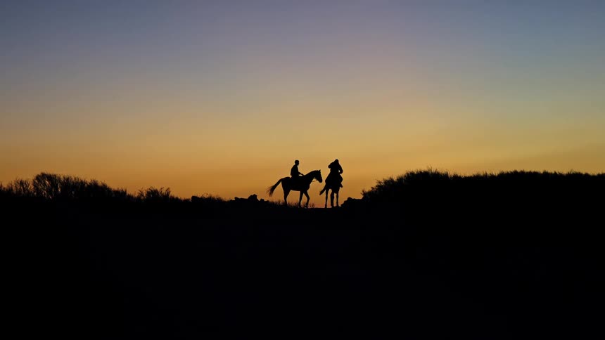 Silhouettes of two riders and their horses stand on a ridge against a gradient sunset sky