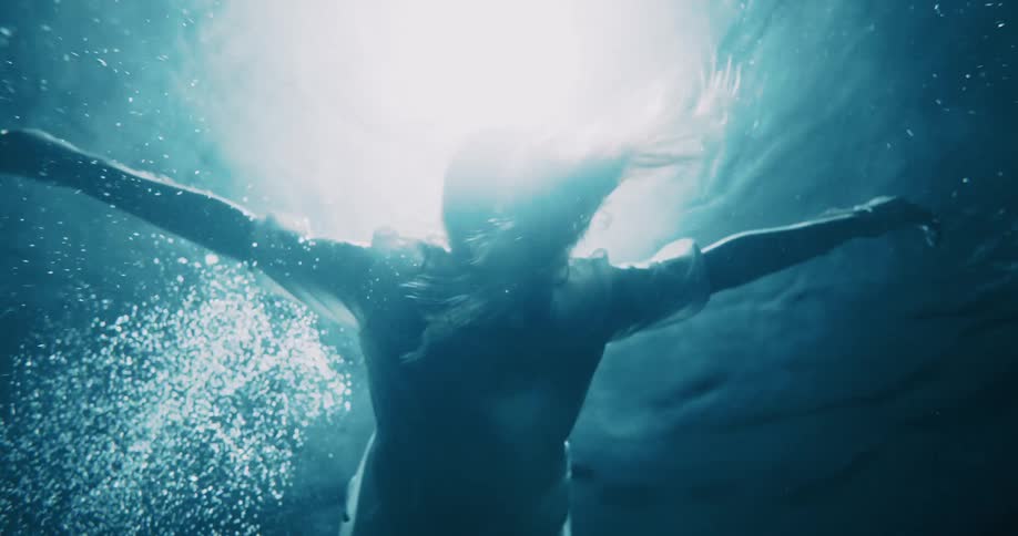 A person is shown underwater arms extended backlit by a strong overhead light creating a dramatic and ethereal scene