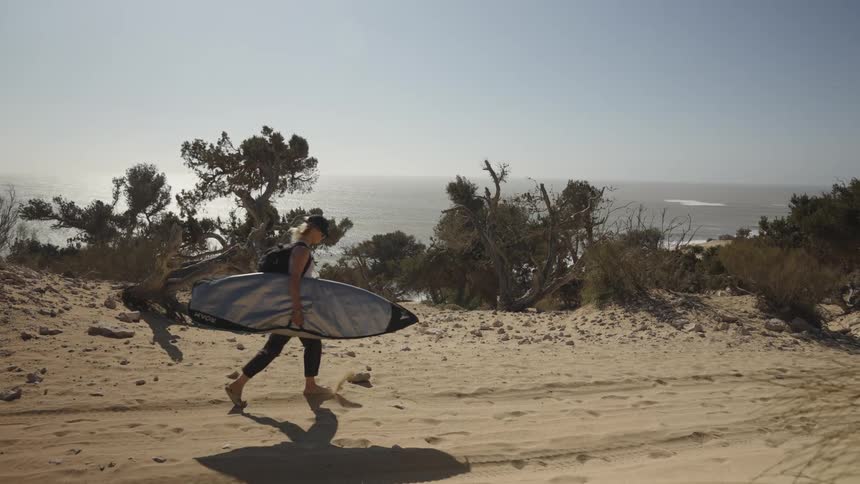 A lady walks towards the beach holding a surfboard on a sunny day