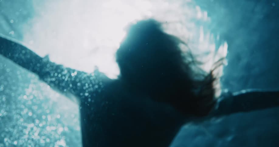 A woman floats submerged underwater her arms outstretched and her hair drifting as beams of light from above create an ethereal glow