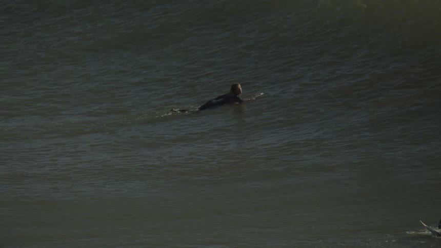 A woman lying on a surfboard paddling and passing through a wave while two other people are surfing nearby