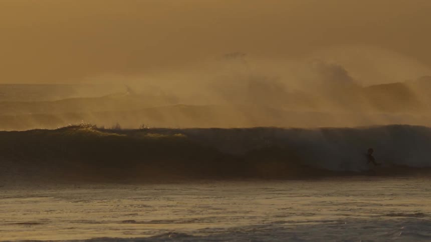 A surfer rides a wave at sunset while others wait in the water silhouetted against the golden horizon
