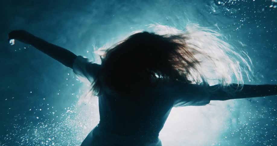 Spotlight shining from above illuminates a woman floating in the water on her back in a dress