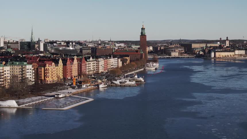 A city skyline featuring a prominent tower overlooks a partially frozen body of water lined with buildings and docked boats