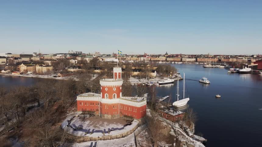 A castle with a fort and a river around it with boats and in the background a city with chimneys and buildings
