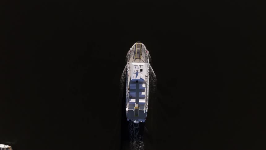 A large white boat sails through dark still water viewed from directly above by a drone