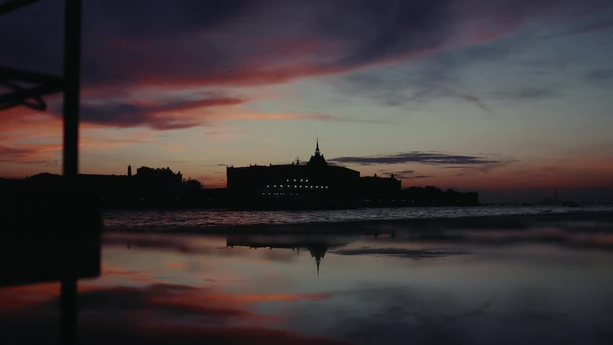 A silhouette of a building with a spire is reflected in calm water during a colorful sunset with hues of orange pink and purple in the sky