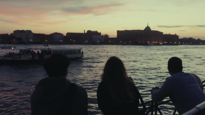 Three individuals stand by a railing gazing at a venetian waterway where boats glide past illuminated buildings under the evening sky
