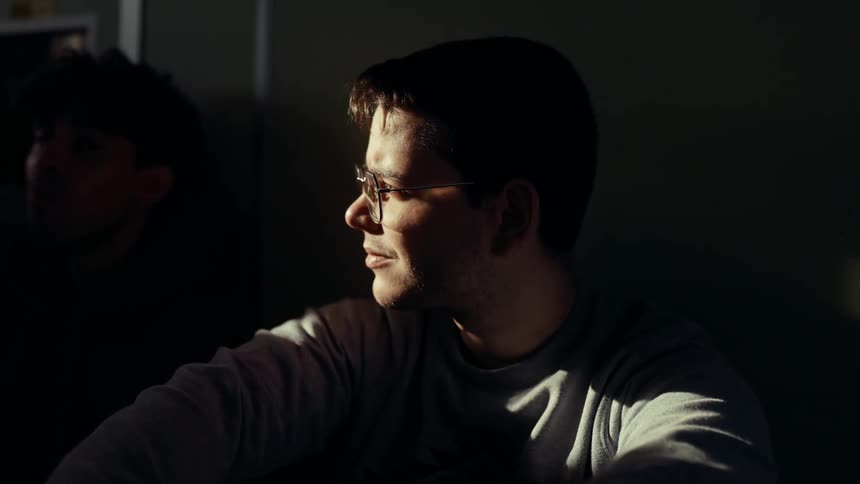 A man wearing glasses is illuminated by strong side light in a dimly lit room