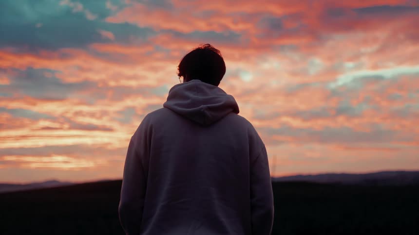 A person wearing a hoodie stands with their back to the camera gazing at a vibrant and colorful sunset sky
