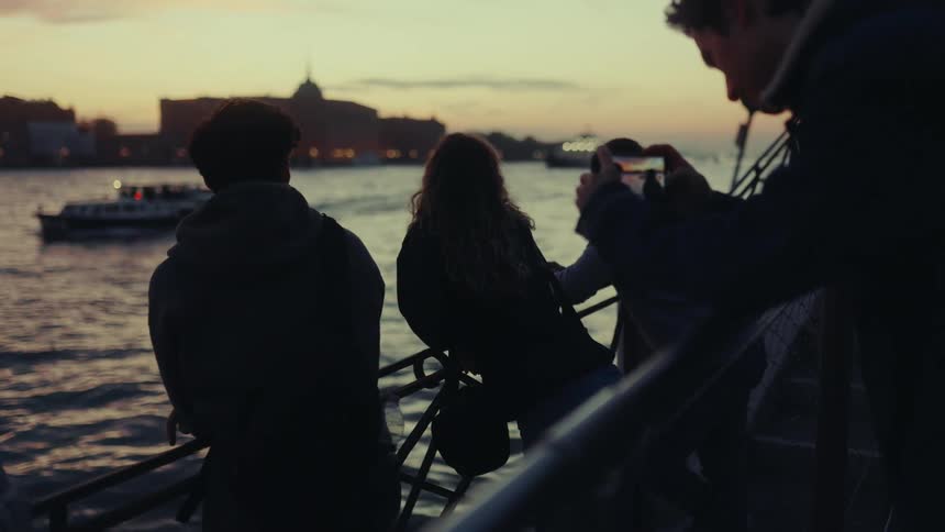 Three people are standing by a waterfront railing at sunset with one person taking a photo of the scene which includes a boat on the water and buildings in the background