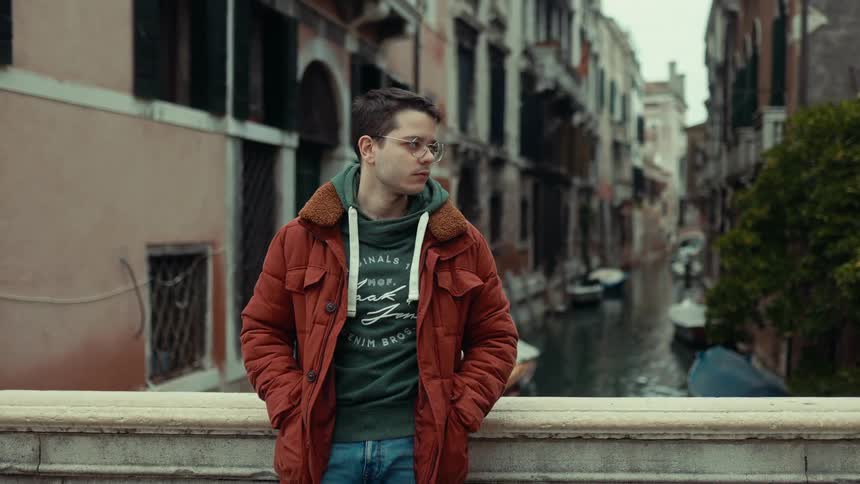 A young man wearing glasses and a jacket stands on a bridge looking to the side as he overlooks a canal lined with buildings