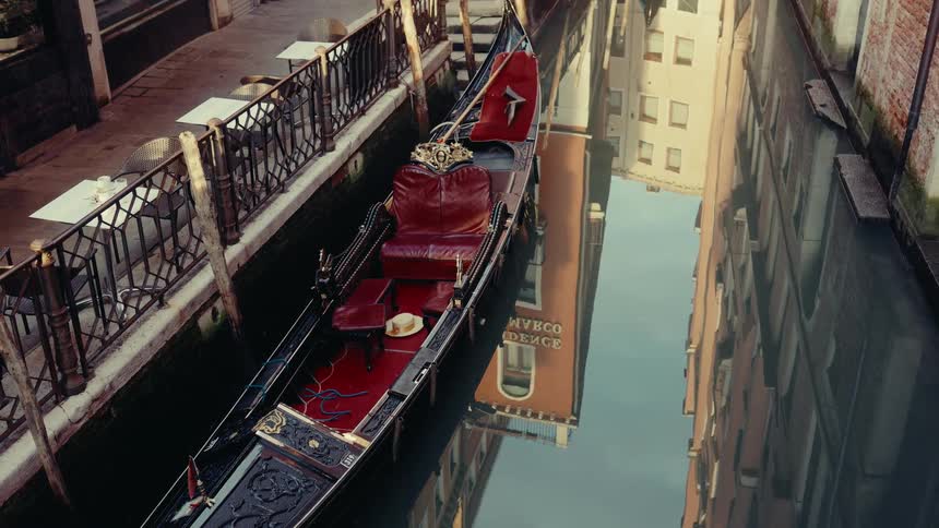 A gondola with red seats floats in a narrow canal reflecting old european buildings in the still water