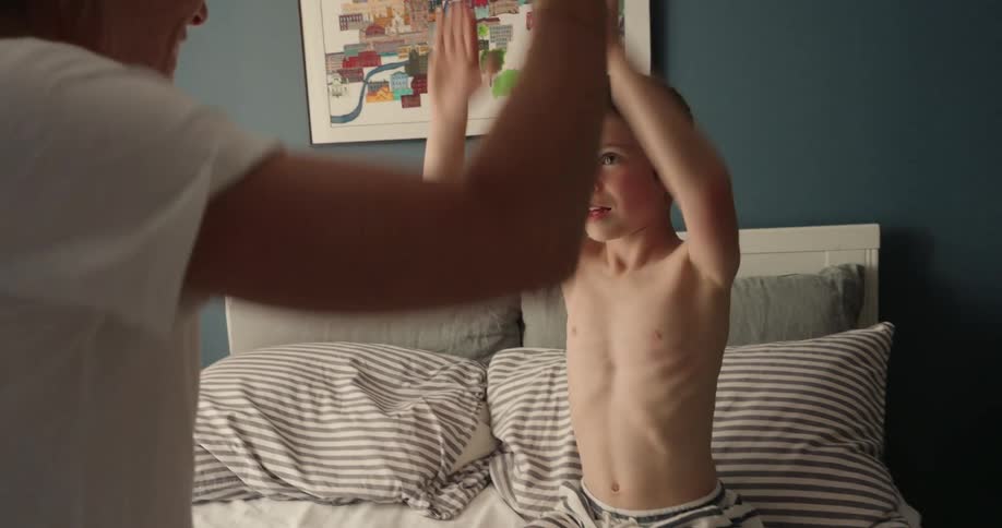 A blondehaired woman with a ponytail joyfully plays with a laughing shirtless child as they sit together on the bed