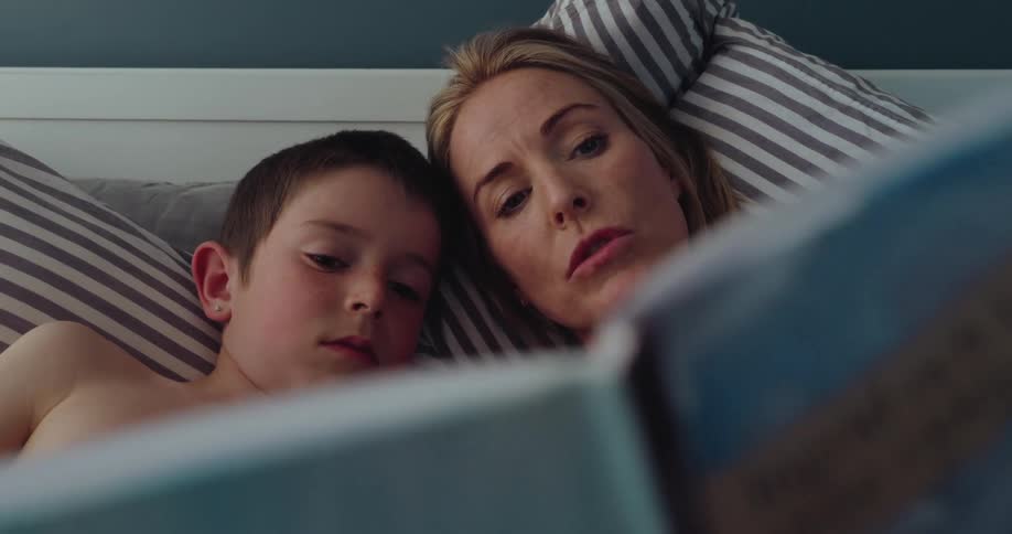 A mother and her son lie together on the bed their heads close as she reads him a book