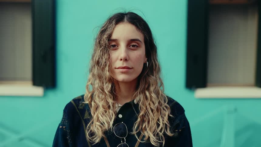 A young woman with wavy blonde hair faces the camera with a neutral expression standing against a bright turquoise wall